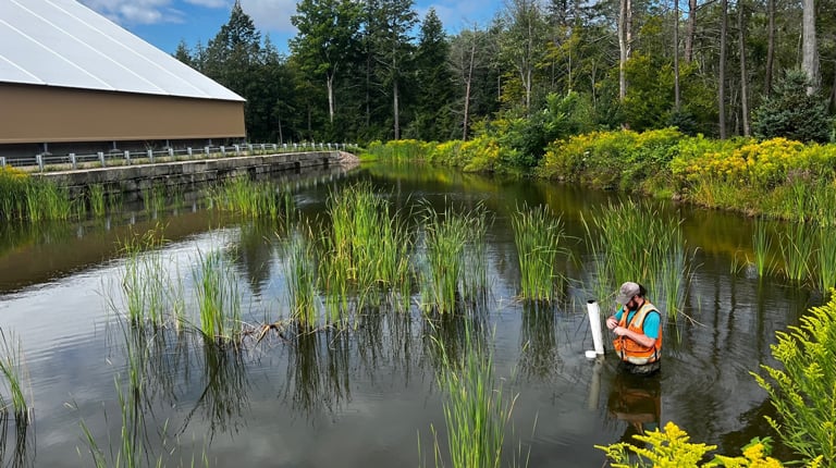 A person in a safety vest conducts water quality testing in a pond surrounded by lush greenery and wetland plants under a partly cloudy sky. A modern building is visible in the background.