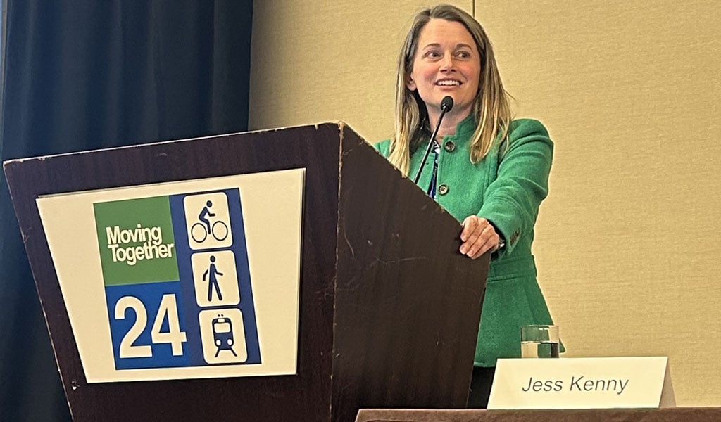 Jess Kenny stands at a podium that reads “Moving Together ‘24” while speaking and wearing business attire.