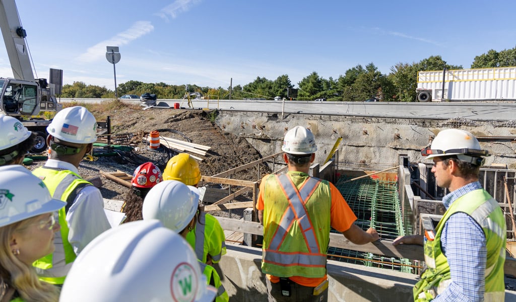 Group of construction workers and students wearing hard hats and reflective vests examining the I95/I90 construction site with heavy equipment and ongoing work in the background.