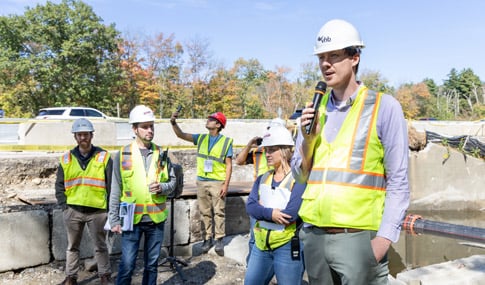 VHB Team Member speaking in a microphone to a group of students wearing construction vests and hard hats with construction machinery in the background. 