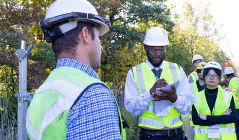 Two students, one taking notes, having a discussion with other workers observing in the background, all wearing safety gear and vests.