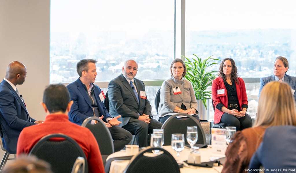 Professional panel discussion at a corporate event, featuring five panelists seated in front of an audience with cityscape views in the background.