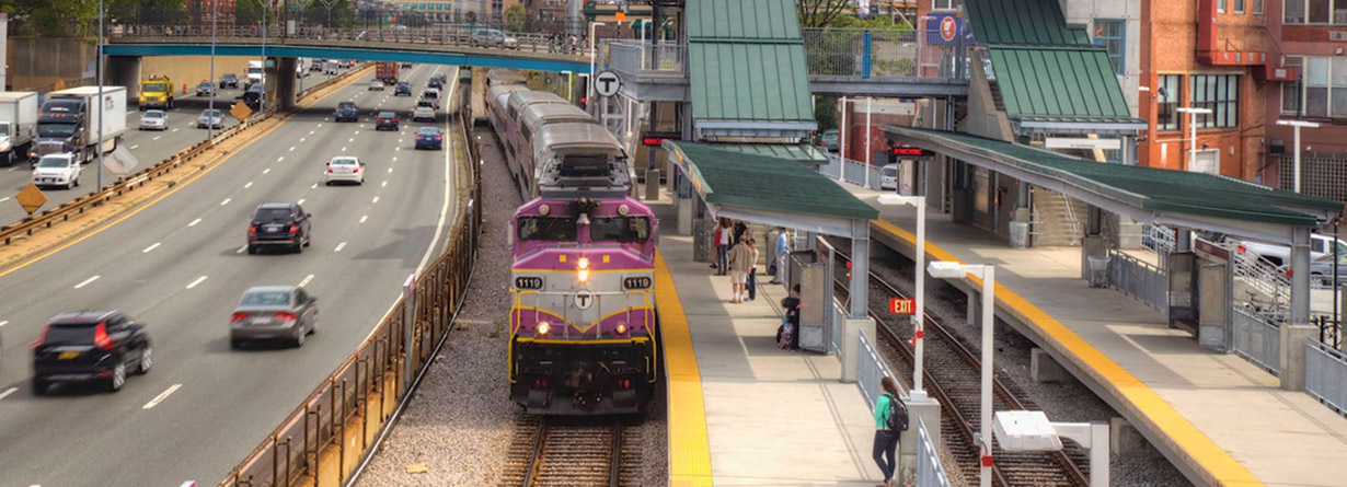 Commuters wait on a platform as a train pulls into the station, with the City of Boston in the background.