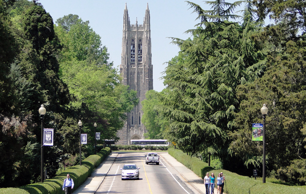 The road leading into Duke University, with people walking on sidewalks and the campus in the background.